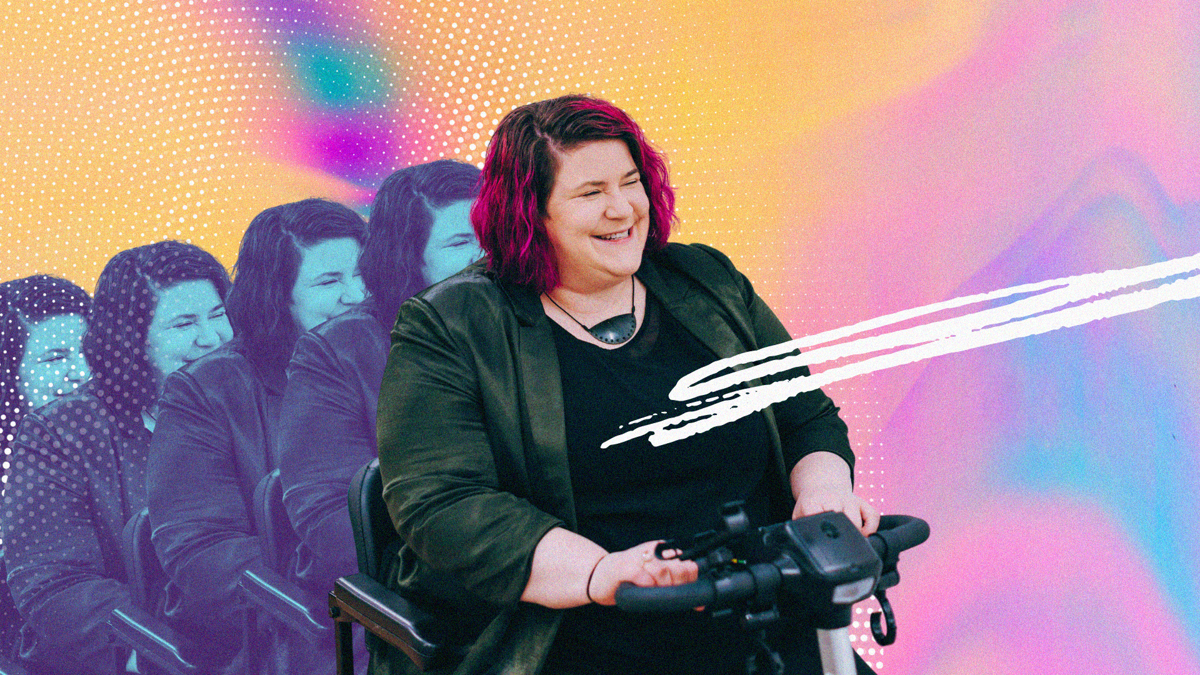 Prudence Walker has pink hair and wears a black jacket and shirt; she's seated in a mobility scooter and grins widely. It looks like she's moving forward at pace with a pink, blue and yellow background behind her.