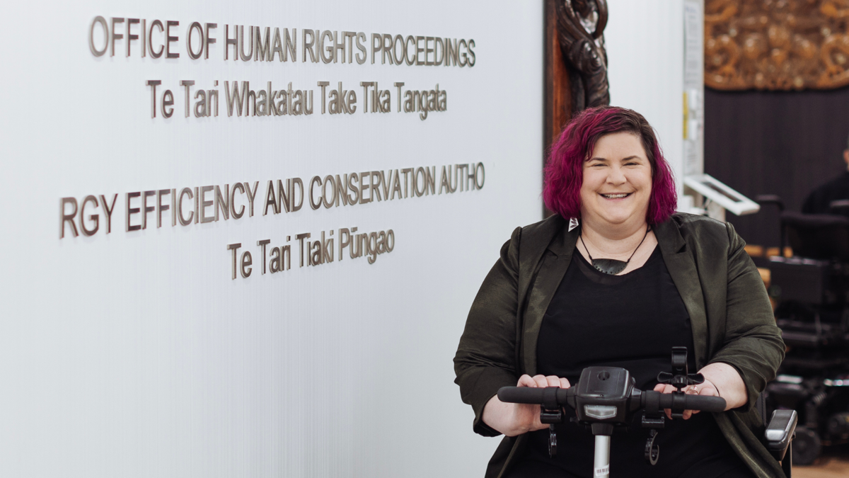 Prudence Walker has pink hair and wears a black jacket and shirt; she's seated in a mobility scooter in front of a wall that reads Office of Human Rights Proceedings. 