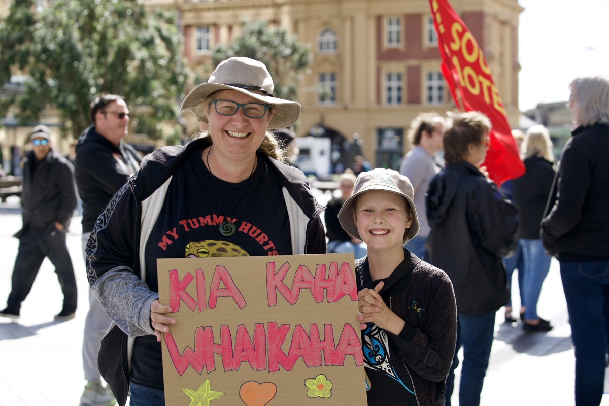 Nicola and a child are smiling as they hold up a cardboard placard that reads “KIA KAHA WHAIKAHA” above a star, heart and flower in fluorescent colours. Nicola’s shirt reads “MY TUMMY HURTS”. 