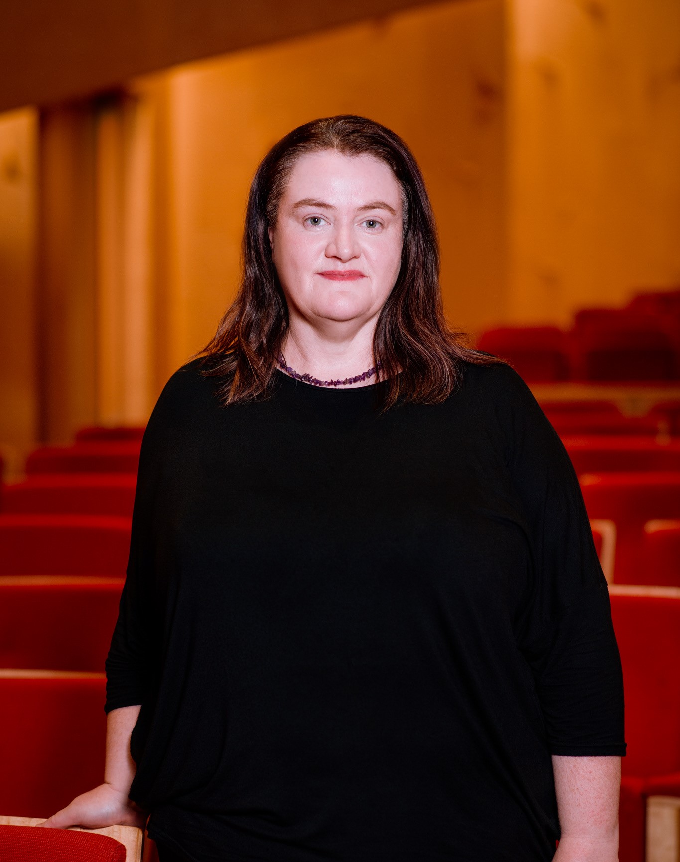 Portrait of Bernie Haldane dressed in black standing among red chairs in a theatre