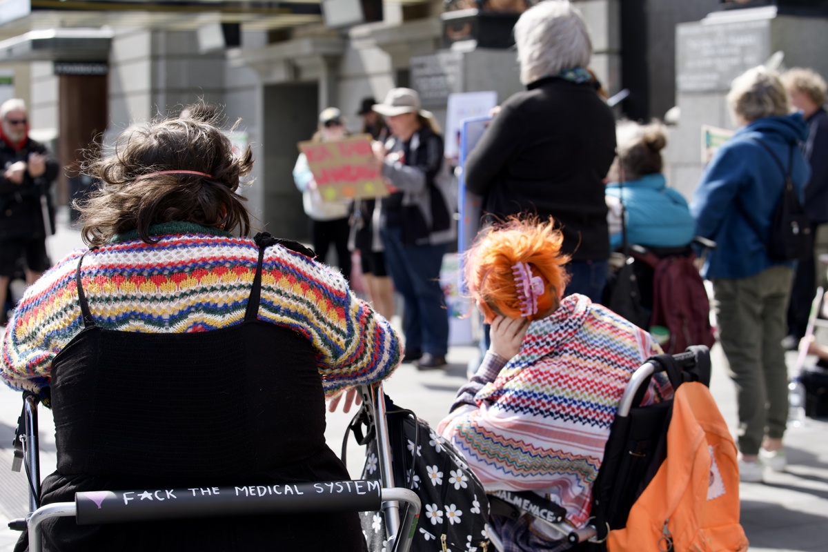  The back of Jazz, seated in a walker, and Finn seated in a wheelchair. Both are wearing colourful knitted clothing and have slogans written on their mobility aids. In the foreground, Jazz’s walker reads “F*CK THE MEDICAL SYSTEM” in white lettering.