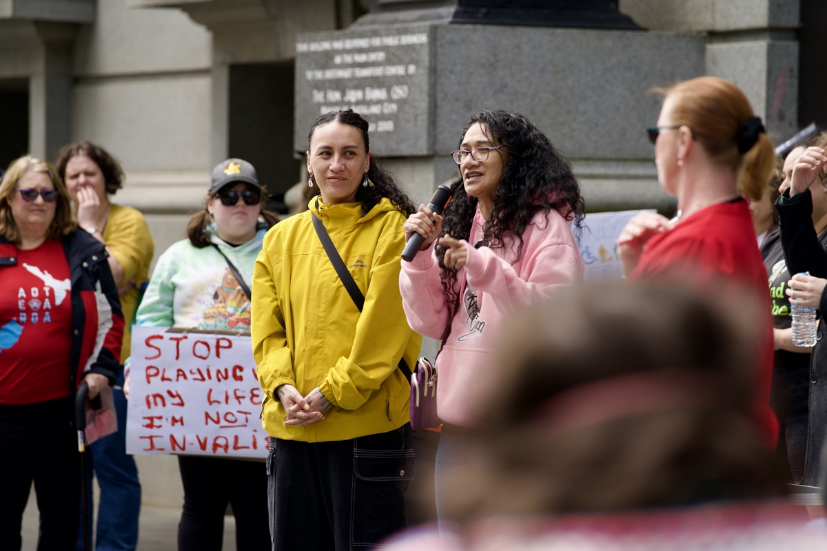 Agnes Magele, who is wearing a light pink hoodie points a finger while speaking while addressing the rally. Brooke Pao Stanley, wearing a bright yellow rain jacket, stands next to Magele. 