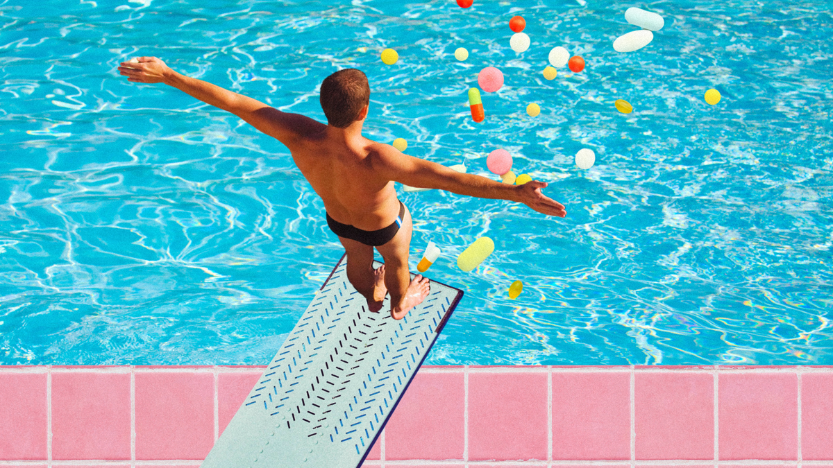 An adult male stands on a diving board about to jump into a pool with colourful pills.