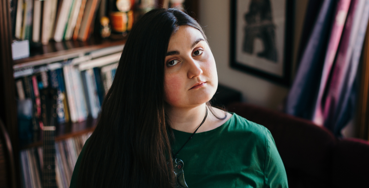 Ruby has long, dark hair and stares into the camera. She wears a dark, green top and green Hei Tiki pounamu. 